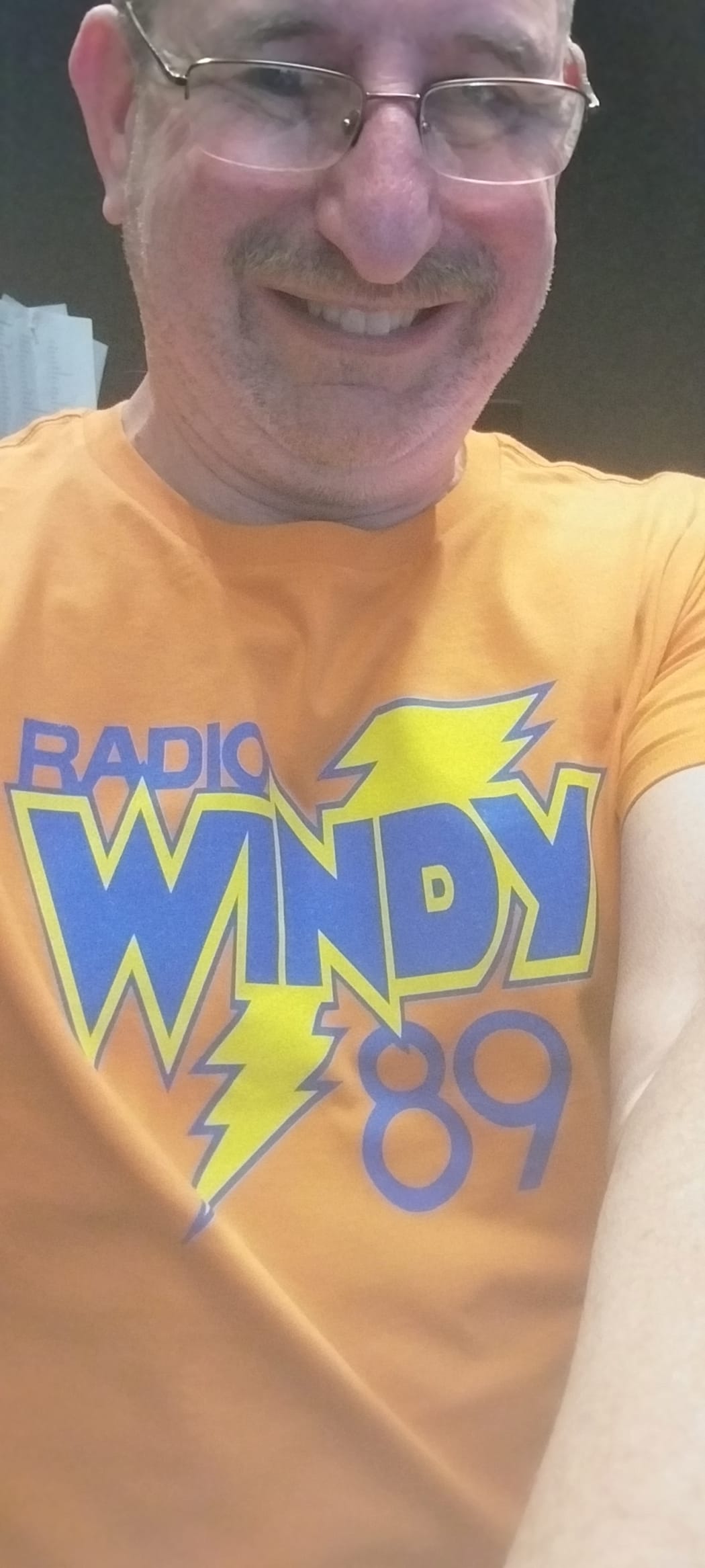 Todd Zaner smiles at the camera. He is wearing an orange tshirt with " RADIO WINDY 89 " written on it.