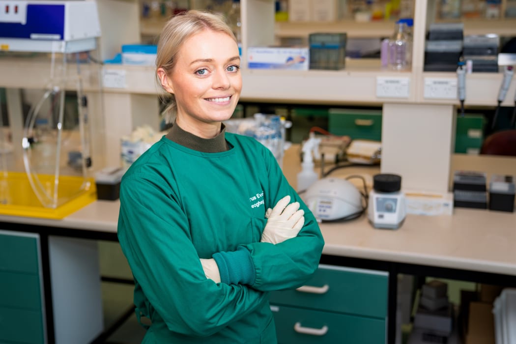 Jemma is smiling and standing with her arms folded in a lab wearing green scrubs and latex gloves