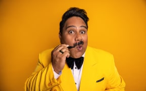 Rutene poses for the camera against a bright yellow backdrop. He wears a yellow suit with a black bowtie, and is drawing on a moustache with a black sharpie.