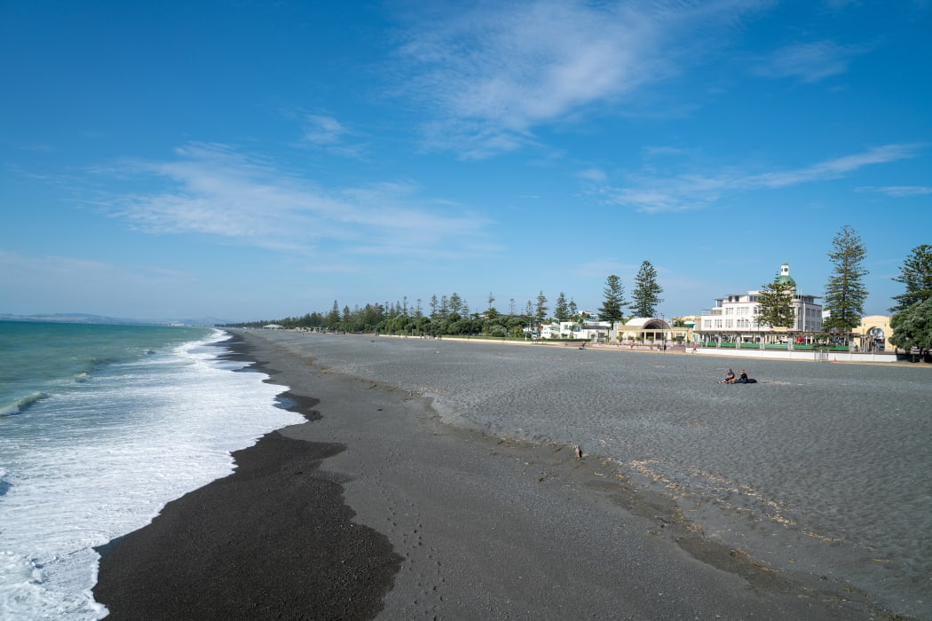 A view along the gray stony Pacific Beach with walkway, Marine Parade Gardens and the soundshell, with Napier city buildings in background.