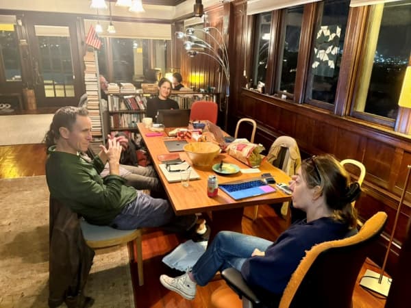 A group of adults chatting at a table, each with paper work and their laptops out.