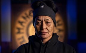 A headshot of April in a black jumper and dark blue beanie in front of an illuminated gang patch on the wall behind her.