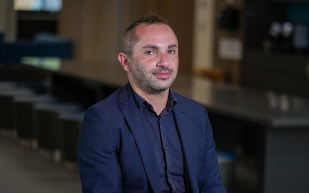 A man with short hair and a short beard sitting in a room. He is wearing a navy shirt and jacket.