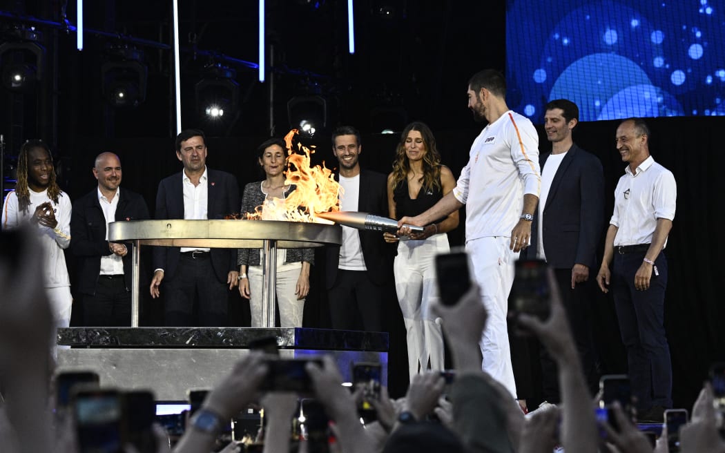 French handball player Nikola Karabati holds the Olympic flame torch to light the cauldron next to France's Minister for Sports and Olympics Amelie Oudea-Castera and President of the Paris 2024 Olympics and Paralympics Organising Committee Tony Estanguet as part of the Olympic torch relay at Place de la Republique in Paris on 15 July 2024, ahead of the Paris 2024 Olympic Games.