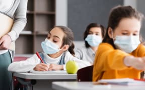 schoolgirl in medical mask near teacher, apple and classmates on blurred foreground