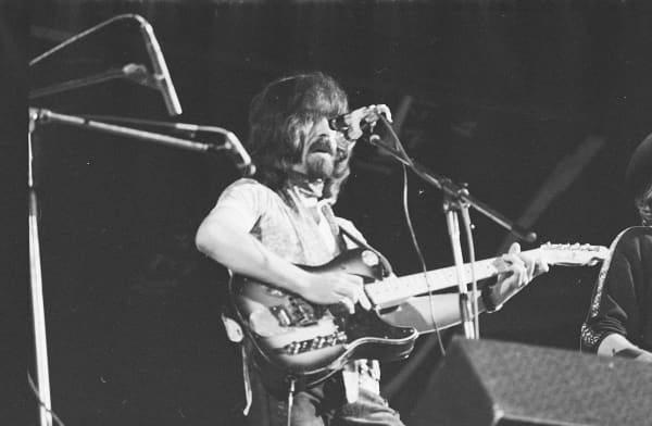 Clarence White - a bearded man with long hair - plays an electric guitar behind a microphone.