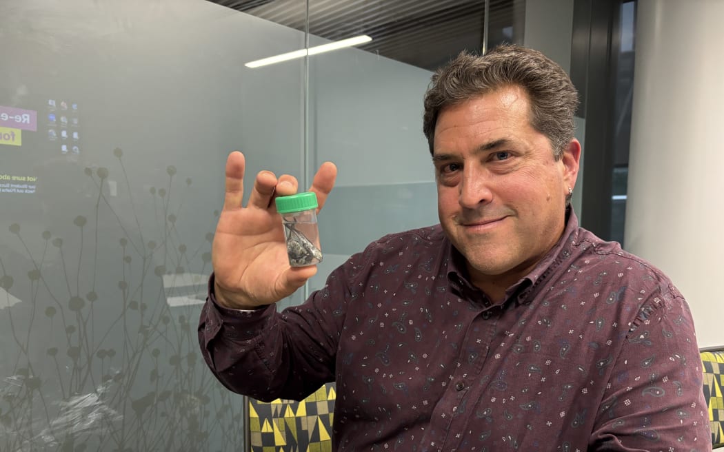 A man in a purple shirt with a grey pattern holds up a small plastic vial with a green lid. Inside the vial are around six silvery fish specimens. The man is looking at the camera and smiling.