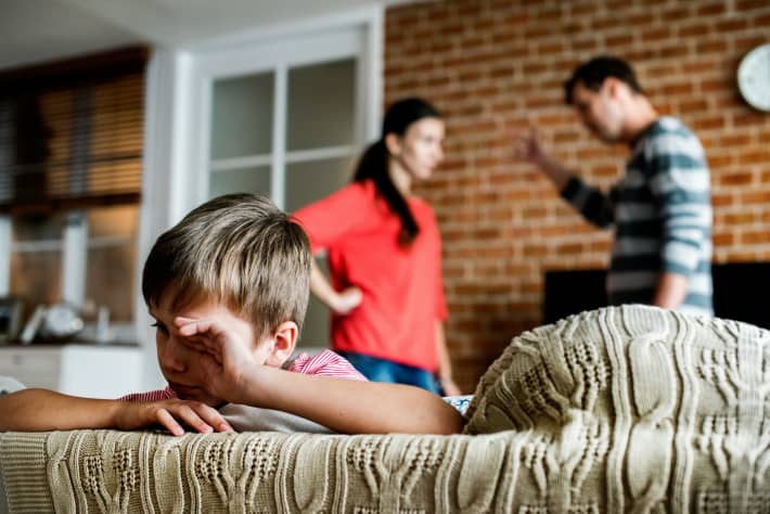 Parenting arguing in background with child wiping tears on sofa.