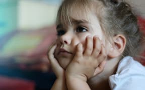 A photo of a little girl lying on the sofa with hands on chin watching the television and looking worried