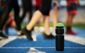 Close up black sports bottle with green cap stands on a blue rubber treadmill with a white line. Water sports drink on the background of a running track with a blue rubber coating with a white line.