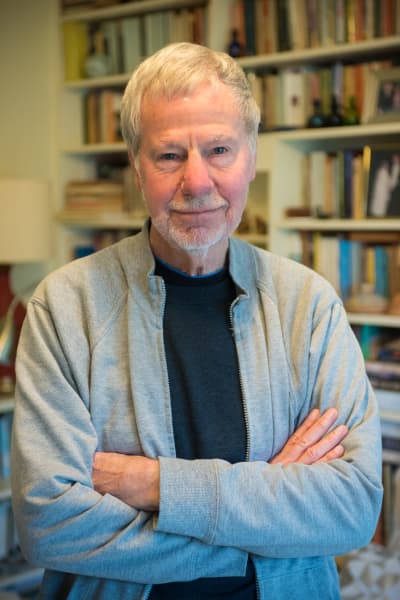 David Hill, a Pakeha man, stands in front of a bookcase. David has grey hair and is wearing a black t-shirt beneath a grey zipped jacket.