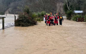 Two people are guided across dangerous floodwaters in Tasman on Friday 11 July, 2025, by members from Fire and Emergency NZ's specialist water response teams from Christchurch and Nelson, using long poles to test what lies under the water.