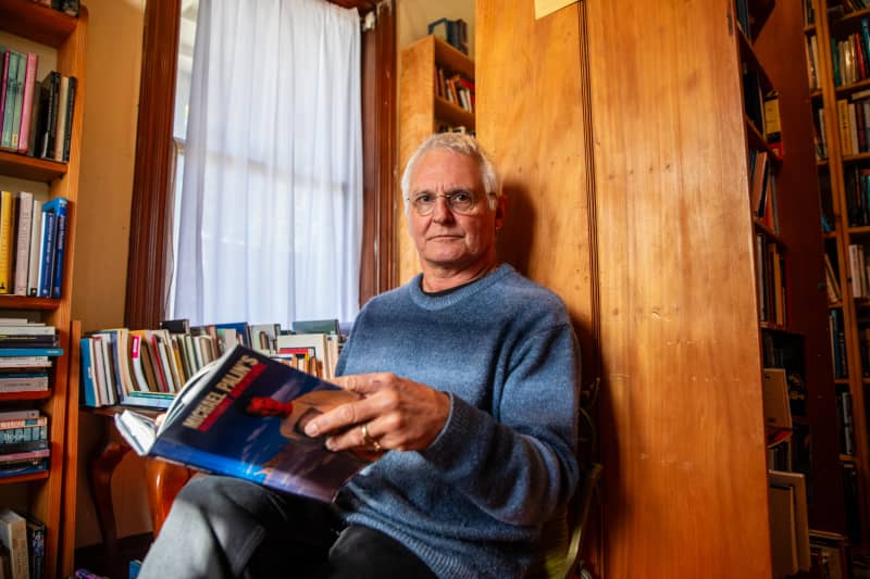 Hard to Find Books shop owner Warwick Jordan sits on a small chair in his shop, with a book open in his hands.