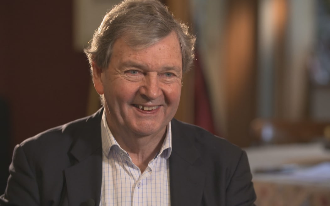 Charles smiles at an interviewer off-screen. He wears a collared shirt and a grey blazer.