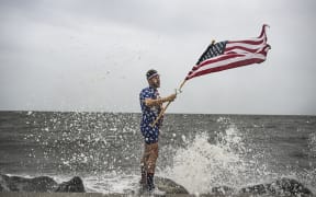 Youtuber Mark Peyton holds a US flag as he poses for his brother Matt Peyton on the shoreline ahead of the arrival of Hurricane Helene in Alligator Point, Florida, on September 26, 2024. Parts of Florida face "unsurvivable" conditions when Hurricane Helene hits later Thursday, the US weather service said, warning that howling wind will drive destructive waves and storm surge as high as 20 feet (six meters) onto the low-lying coast. Residents heeded mass evacuation orders and fled ahead of the incoming hurricane -- projected to be one of the largest Gulf of Mexico storms in decades. (Photo by CHANDAN KHANNA / AFP)