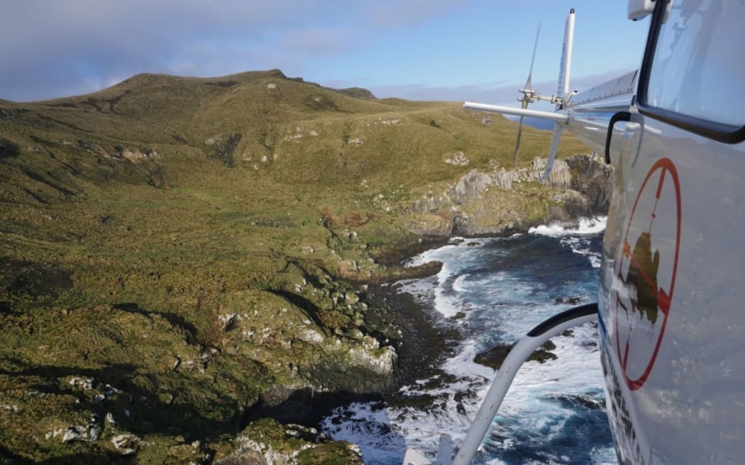 An aerial shot of rugged cliffs and wild seas as viewed from a helicopter. The tail and side of the helicopter, featuring a logo of a mouse silhouette in a red target, is visible on the right of the image.