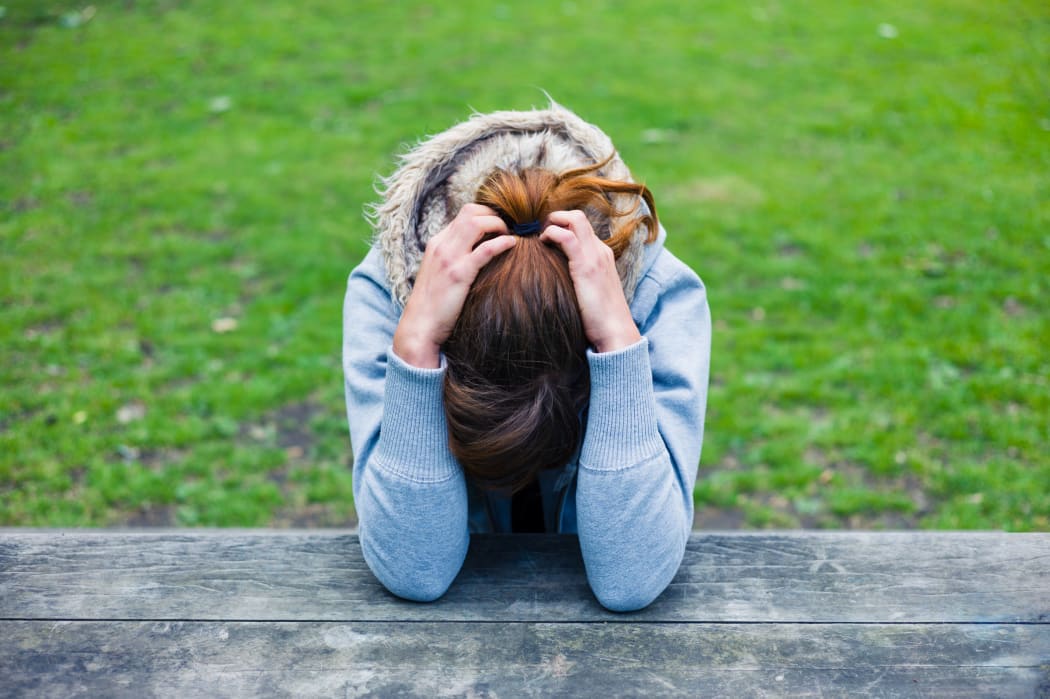 A young woman - sad or upset - sits on a bench with her head in her hands.