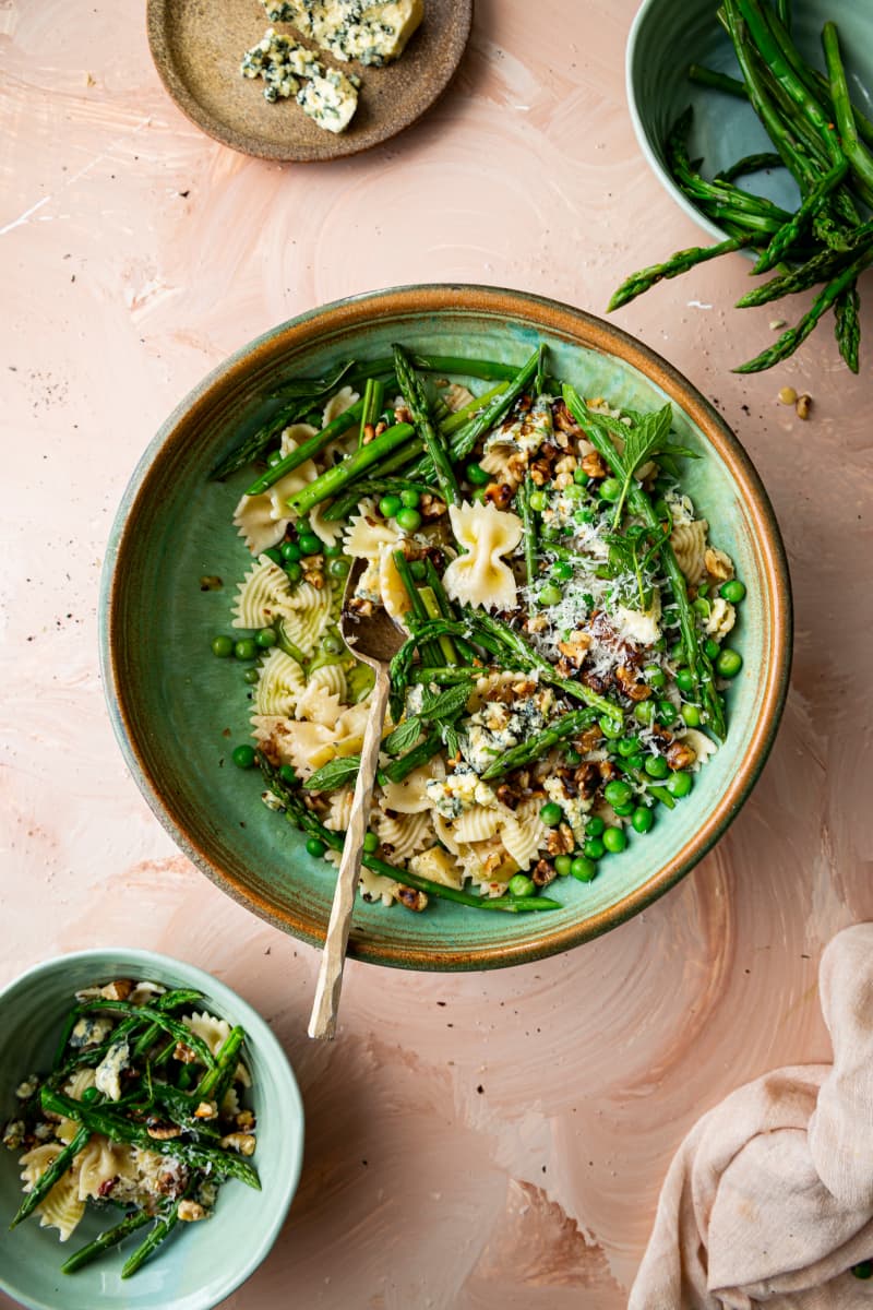 A bowl of farfalle (bow-shaped) pasta with asparagus, peas, blue cheese and walnuts on a pale pink background.