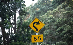 A road sign indicating a 65km speed around a tight corner on a rural road in the Coromandel.
