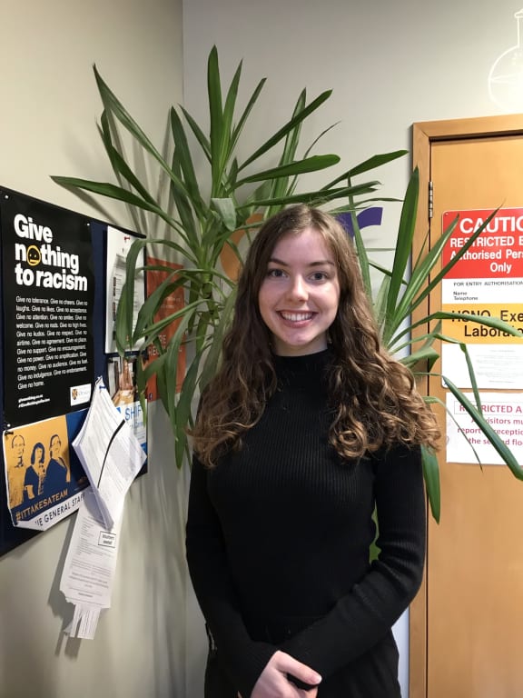 A photograph of Jemma Ellie taken in front of a plant in the foyer of the Biochemistry Department.