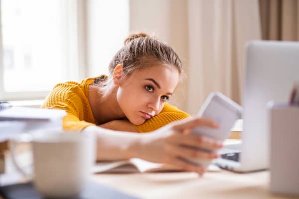 A bored-looking girl looks at her phone as she leans on desk with laptop open.