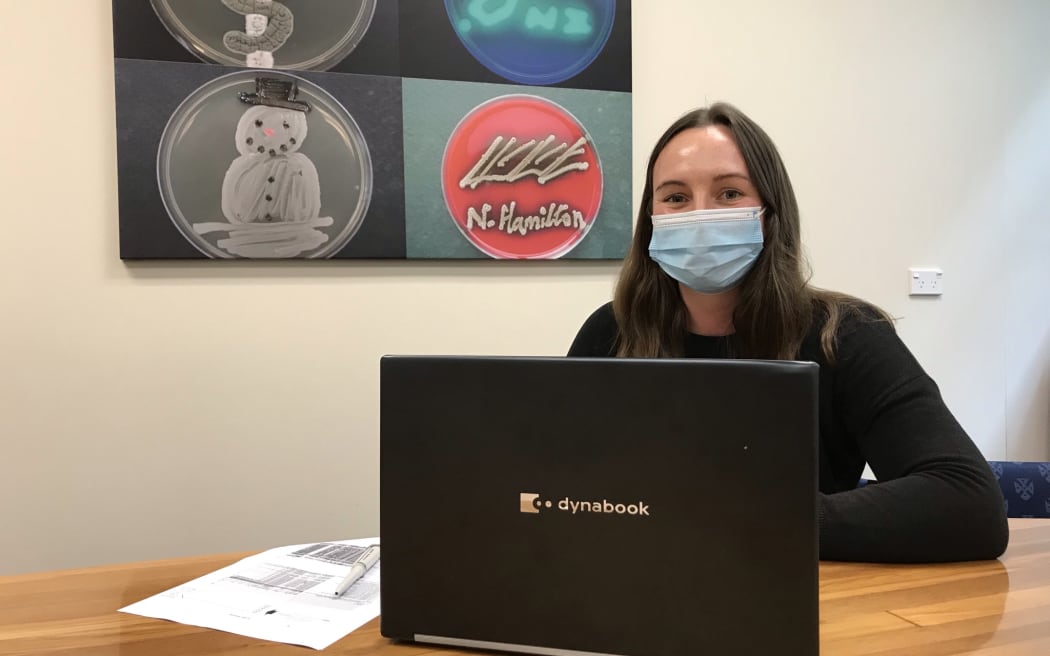Stephanie is sitting at a table behind a laptop. She is wearing a face mask.
