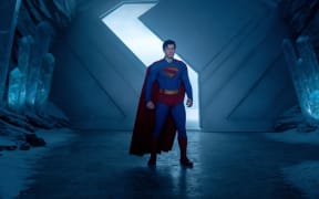 A still from the 2025 movie Superman. Superman stands in the entrance of a great icy cave, wearing his red cape and supersuit.