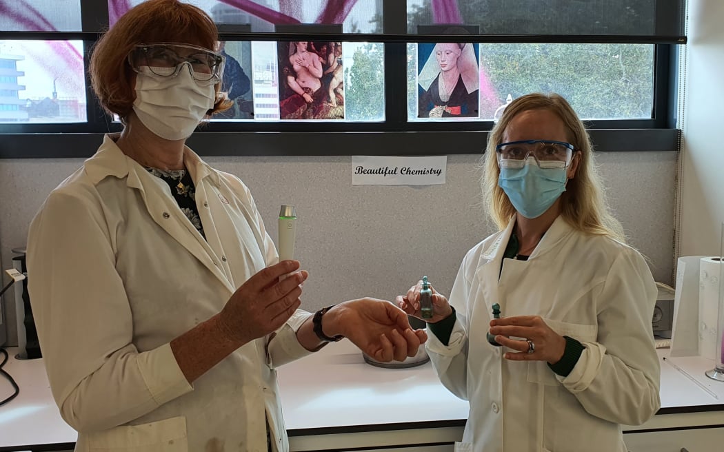 Michel and Erin stand before the Beautiful Chemistry lab space. Erin holds two small glass vials with extract in them. Michel holds an instrument to measure skin moisture. They are both wearing lab coats, goggles and face masks. There are Renaissance images on the window behind them.