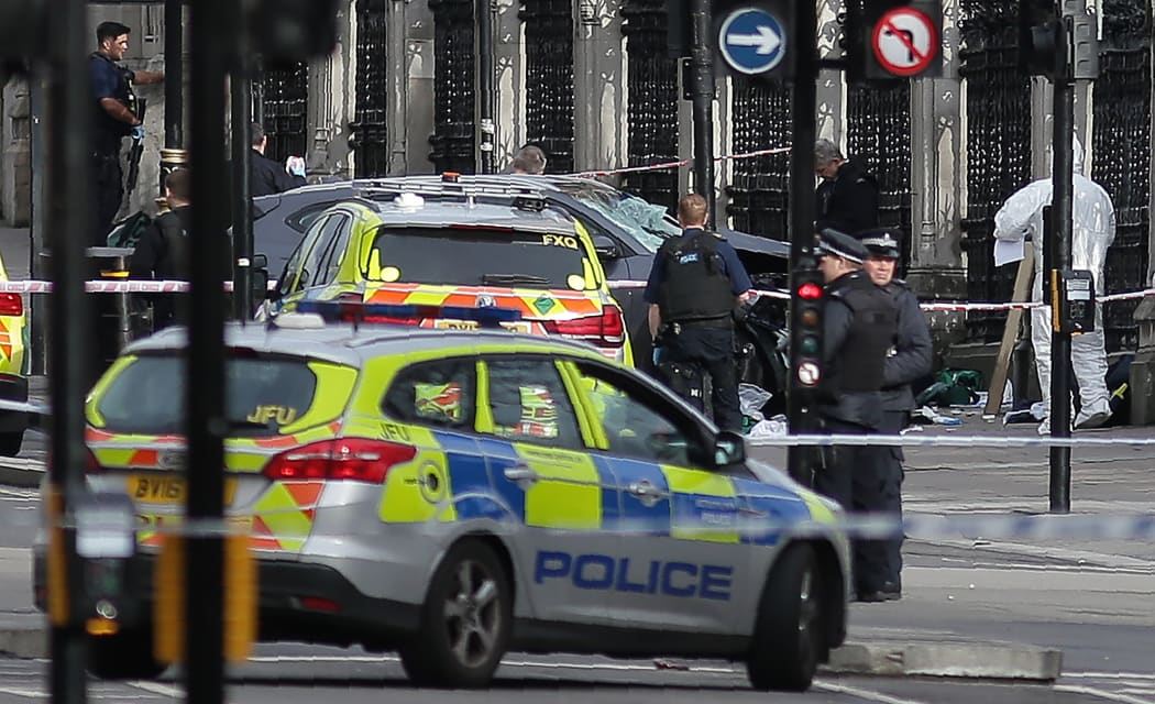 Armed police officers stand guard, as forensics officers work around a grey vehicle that crashed into the railings of the Houses of Parliament during an emergency incident.