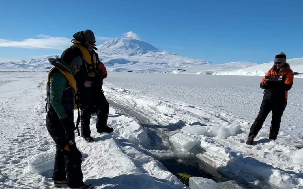 Three people gather around a crack in the sea ice with a snowy mountain visible in the distance. The sky is blue. One person, on the right of the crack, is wearing a classic navy and orange Antarctica New Zealand jacket and a beanie. The other two people, on the left of the crack, are wearing beanies, dry-suit waders, and lifejackets.
