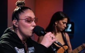 Theia is singing into a microphone. There is a bass player in soft focus behind her. In the background you can see the Z of some RNZ branding.