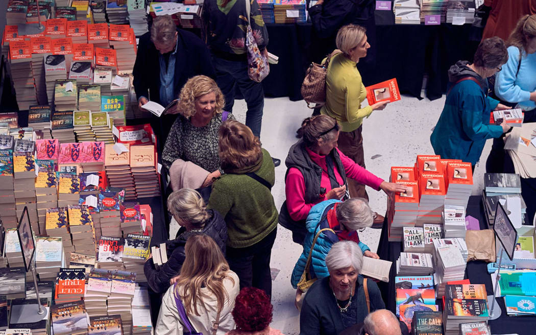 An overhead shot of a large group of people milling between tables stacked high with books. People browse books, chat to one another, and make purchases.