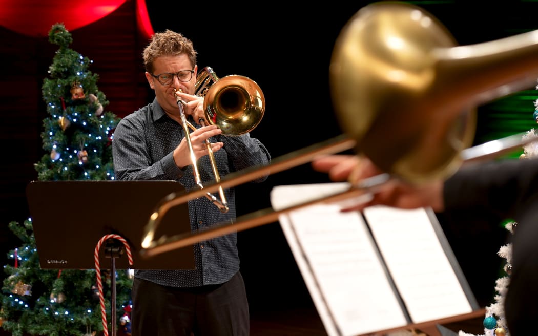 Matthew Allison plays his trombone in front of a Christmas tree. The foreground features another trombone and a music stand.