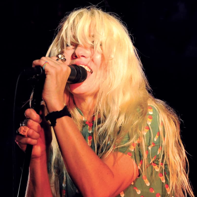 A woman with long blonde hair concealing her eyes sings into a microphone.