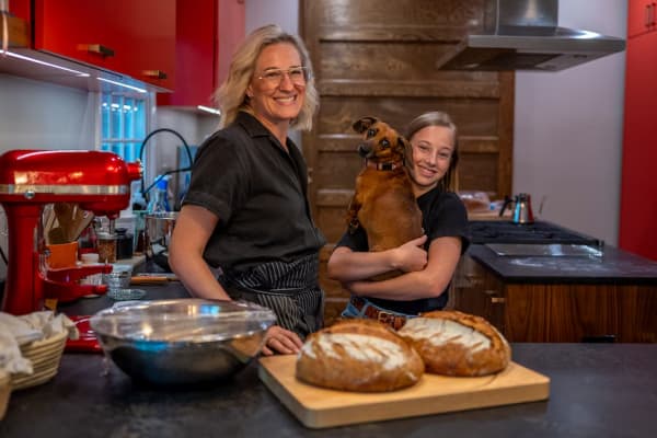 Catherine Pakaluk standing in her kitchen with two loaves on a cutting board, with her daughter Valerie beside her holding their dog Queenie.