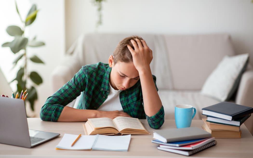 Bored teenager reading book at desk at home, holding his head, free space