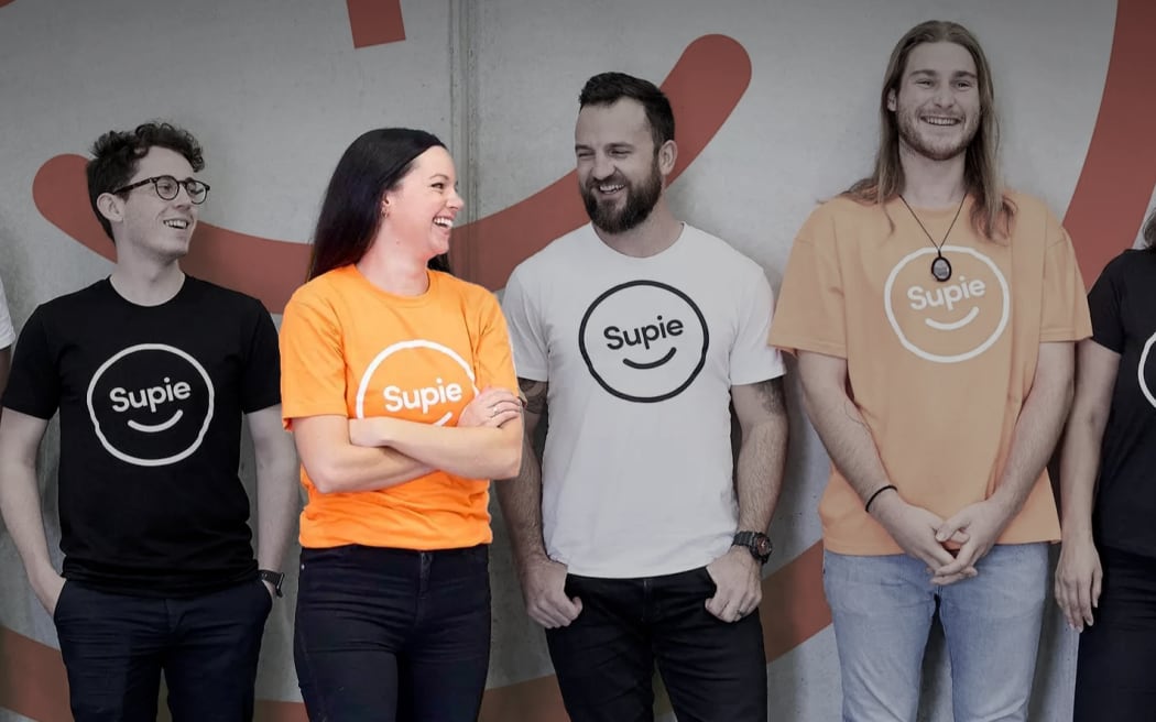 A group of five people smiling and laughing, wearing "SUPIE" t-shirts in black, white and orange. Sarah Balle is spotlit, while the other people are slightly in shadow.