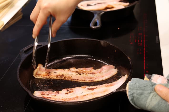 Two slices of bacon being cooked on a frying pan.