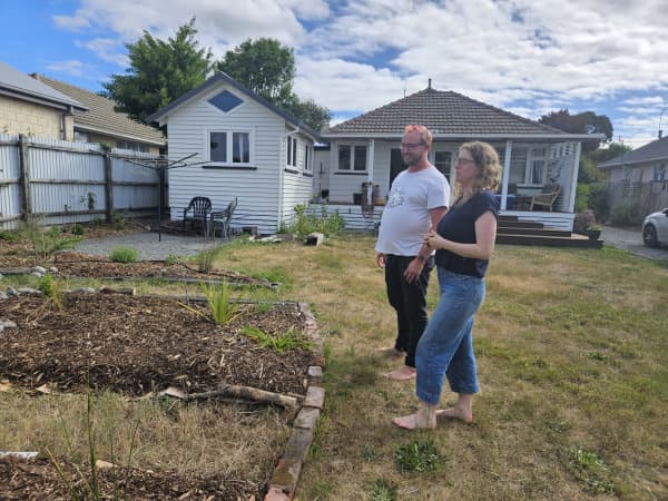 A man and a woman are standing barefoot on a lawn, looking at a garden plot. In the back ground you can see a house and deck.