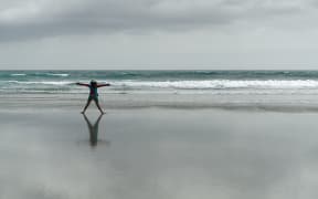 Girl, back to camera, doing exercises stretching arms outstretched on an empty beach, New Zealand.
