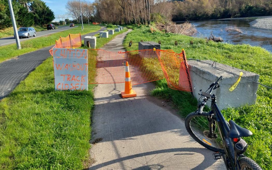 A path beside Te Awa Kairangi is closed by a ‘Caution, washout, track closed’ sign due to recent erosion on the east side of the river. The detour path and Harcourt Werry Dr are to the left.