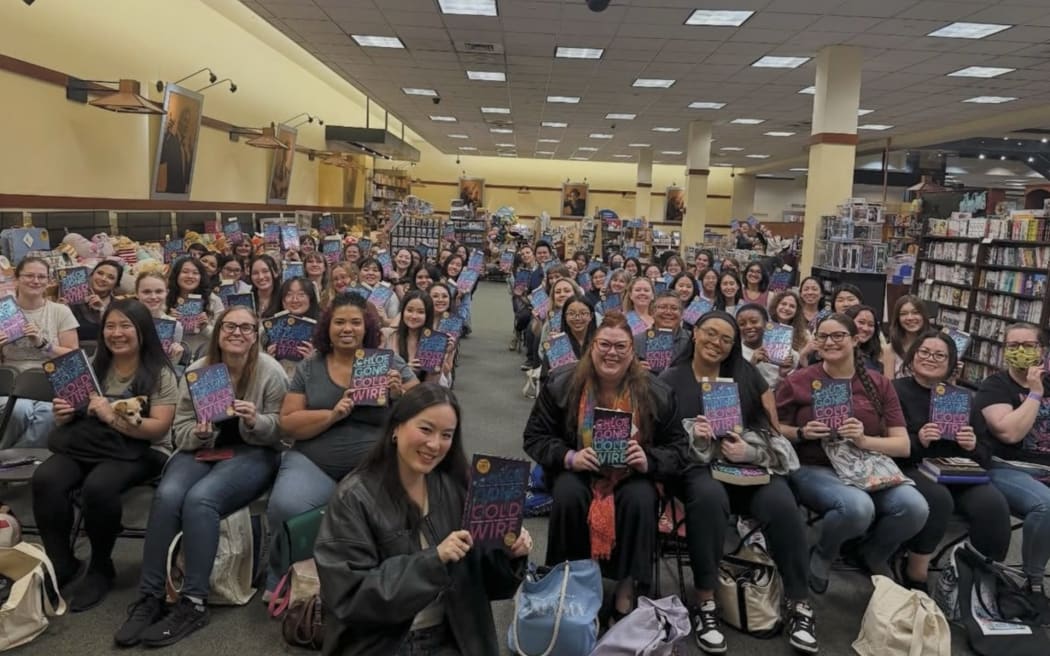 Chloe Gong on her US book tour holds up a copy of her latest book, Cold Wire, as she sits between dozens of fans holding her book in a hall.