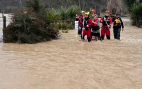 Two people are guided across dangerous floodwaters in Tasman on Friday 11 July, 2025, by members from Fire and Emergency NZ's specialist water response teams from Christchurch and Nelson, using long poles to test what lies under the water.