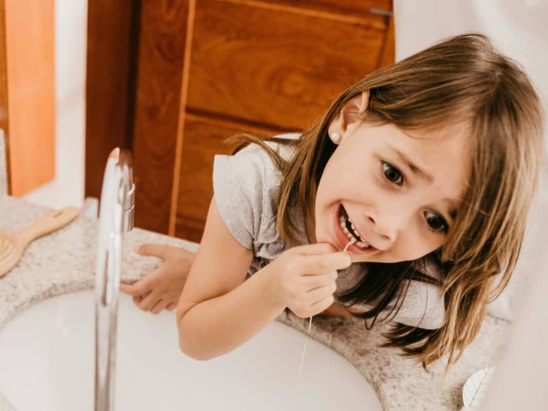 A young girl flosses her teeth at the bathroom sink.