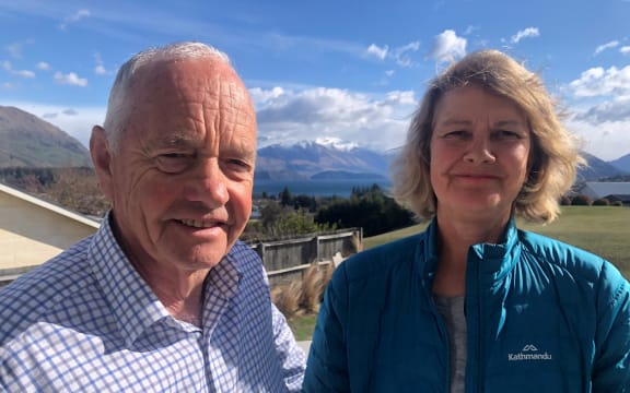 Michael and Meg smile at the camera. Michael wears a navy and white checked shirt, and Meg wears a turquoise outdoors jacket. In the background is Lake Wānaka and snowcapped mountains beneath a blue sky.
