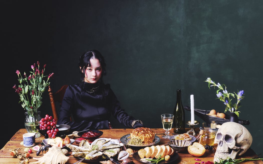 Singer and author Michelle Zauner sits at a table covered in food, a candle and a skull. Michelle wears black.