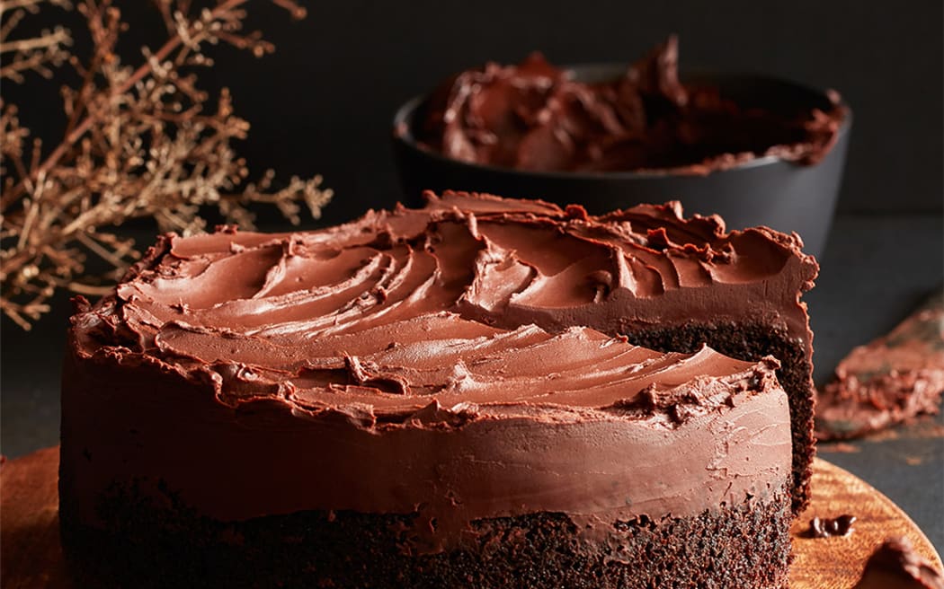 Image of a round chocolate cake with a thick head of chocolate ganache icing. The cake is on a wooden board in the foreground, with a bowl of ganache icing lurking at the back. A slice of cake is visible in the front righthand corner.