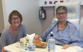 Two people in a hospital setting. One is in a raised bed with a tray in front of them with food and drink. To the left is Treza, smiling and holding the hand of the person in bed.