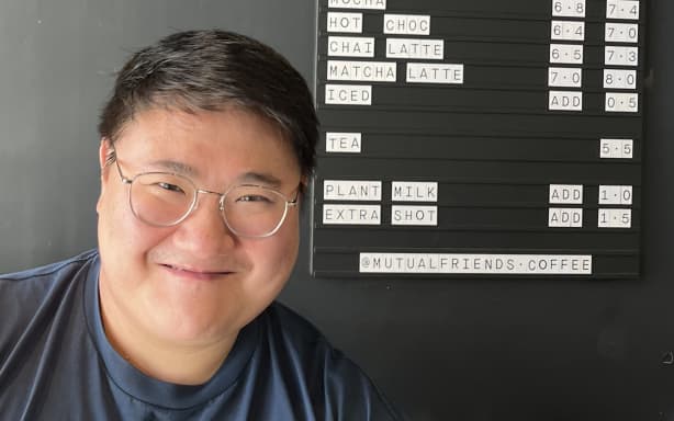A head shot of Hubs Tong, Head Educator at MQ Coffee in front of the menu board of the cafe he co-owns Mutual Friends.