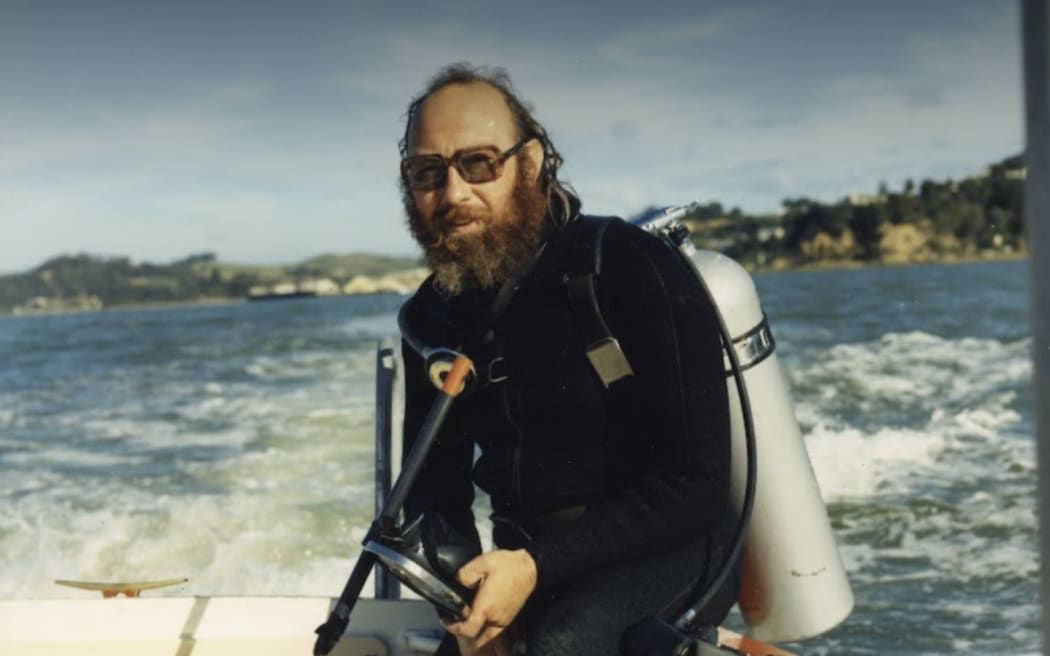 A man with a beard and glasses, dressed in an old-style black wetsuit and scuba gear, sits on the edge of a small boat. He holds a diving mask and looks toward the camera, with choppy water and a distant shoreline visible behind him under a partly cloudy sky.